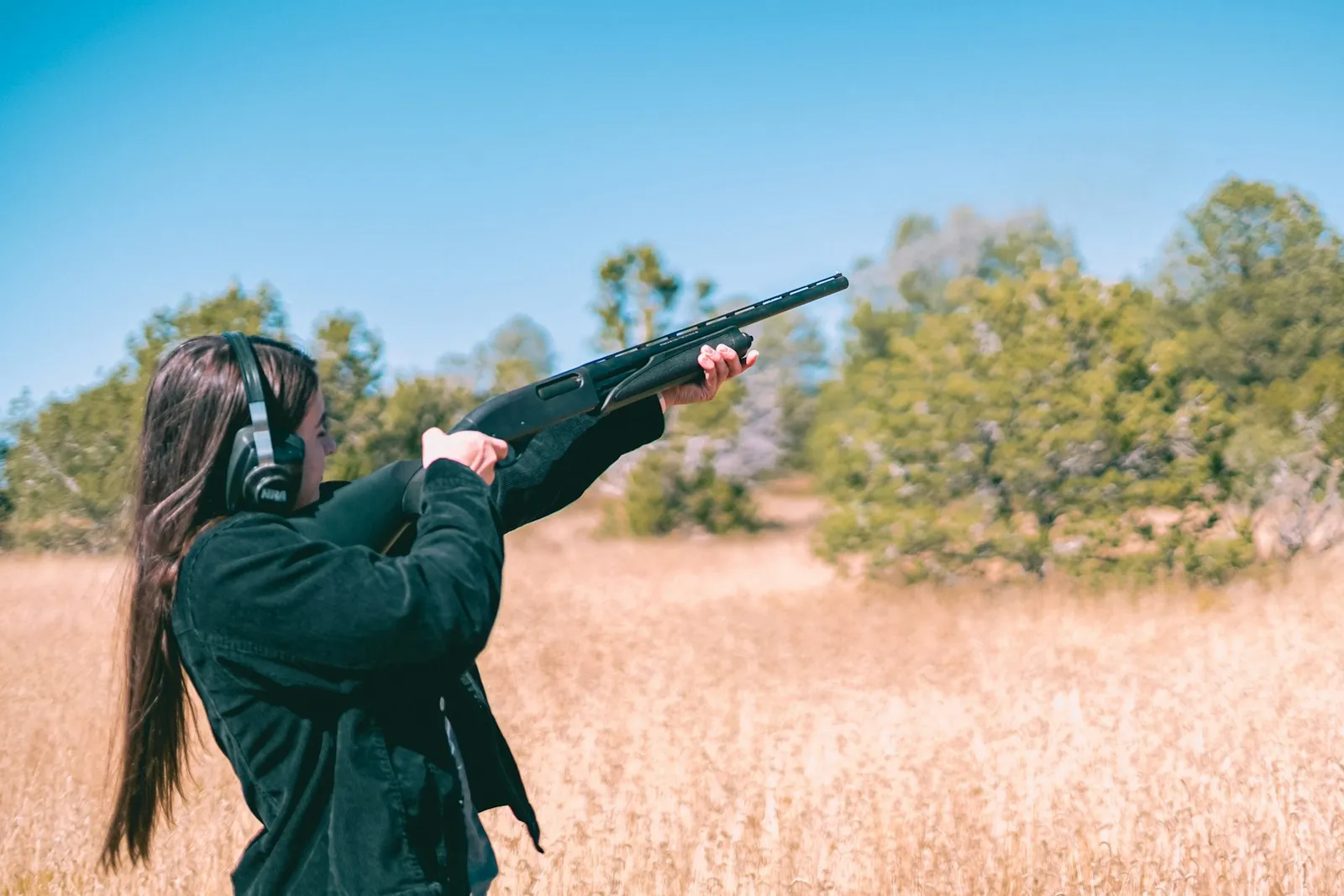 Tireuse avec casque antibruit épaulant un fusil dans un champ de hautes herbes